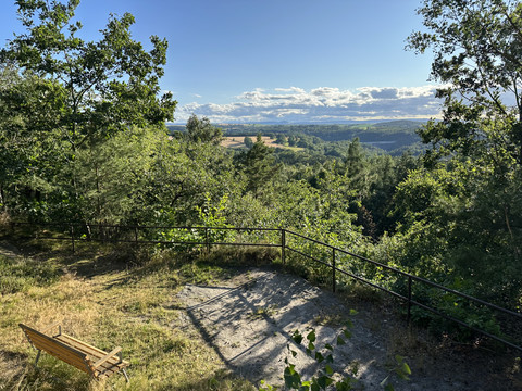 Holzbank auf einem Hügel mit Blick auf eine grüne, bewaldete Landschaft unter blauem Himmel und vereinzelten Wolken.