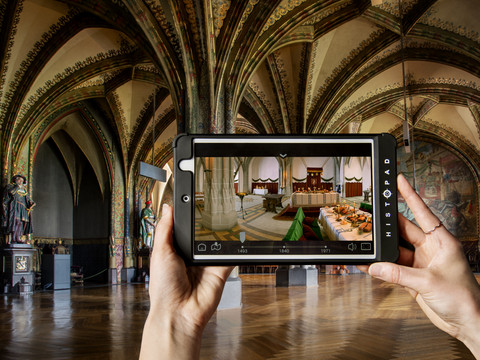 Hände halten ein Tablet mit virtueller Darstellung einer historischen Innenhalle.Hands holding a tablet with a virtual representation of a historic interior hall.Ruce držící tablet s virtuálním vyobrazením historické interiérové haly.Ręce trzymające tablet z wirtualnym odwzorowaniem historycznego wnętrza holu.Handen die een tablet vasthouden met een virtuele voorstelling van een historische binnenhal.Mani che tengono una tavoletta con una rappresentazione virtuale di una sala interna storica.
