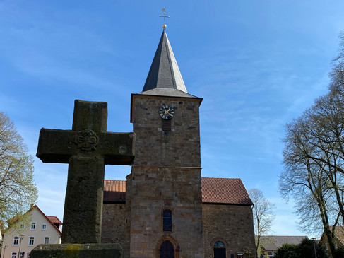 Steinkirche mit Turm und Kreuz, umgeben von Bäumen an einem klaren blauen Himmel.