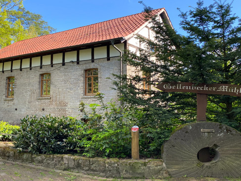 Historische Ziegelmühle mit rotem Dach, umgeben von Bäumen und einem alten Mühlstein im Vordergrund.