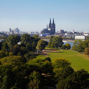Rhine Park Das Bild zeigt den Kölner Rheinpark mit seiner weitläufigen Grünfläche. Im Hintergrund erheben sich majestätisch der Kölner Dom und die Altstadt, flankiert von der Hohenzollernbrücke. Bäume und Büsche säumen die Wiesen.The picture shows Cologne's Rhine Park with its extensive green space. Cologne Cathedral and the old town rise majestically in the background, flanked by the Hohenzollern Bridge. Trees and bushes line the meadows.