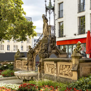 Pixies’ Fountain Heinzelmännchenbrunnen in Köln: Brunnen mit Figuren und Reliefs, umgeben von Blumen.Heinzelmännchenbrunnen in Cologne: fountain with figures and reliefs, surrounded by flowers.