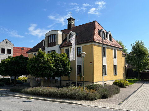 touristinformation-dittersbach-(c)-emily-wolf-CC-BY-(2).jpg Gelbes Gebäude mit rotem Ziegeldach und Uhrturm, umgeben von Bäumen und Sträuchern unter blauem Himmel.Yellow building with red tiled roof and clock tower, surrounded by trees and bushes under a blue sky.Žlutá budova s červenou taškovou střechou a věží s hodinami, obklopená stromy a keři pod modrou oblohou.Żółty budynek z czerwonym dachem pokrytym dachówką i wieżą zegarową, otoczony drzewami i krzewami pod błękitnym niebem.Geel gebouw met rood pannendak en klokkentoren, omringd door bomen en struiken onder een blauwe lucht.Edificio giallo con tetto di tegole rosse e torre dell'orologio, circondato da alberi e cespugli sotto un cielo blu.