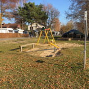 Spielplatz Lüttjen Mardorf Spielplatz mit Schaukel und Sandkasten auf einer sonnigen Wiese, umrahmt von Bäumen und Wohnhäusern.