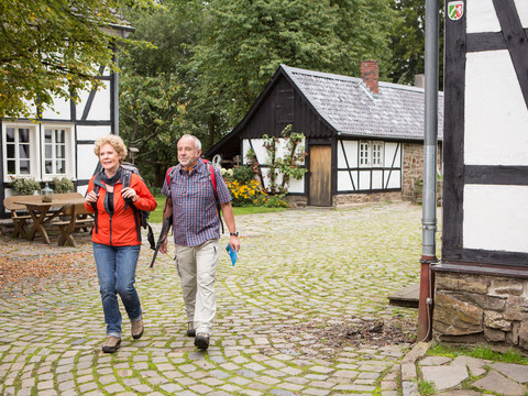 Wanderer am Kochshof <p>Ein älteres Paar wandert auf einem gepflasterten Weg durch ein malerisches Fachwerkdorf.</p>
