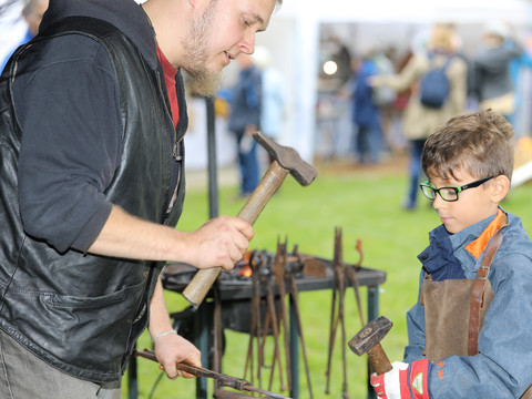 Herbstmarkt Ein Schmied zeigt einem Jungen, wie man mit Hammer und Amboss arbeitet, um Metall zu formen.