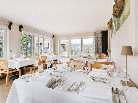 Helles Restaurant mit großen Fenstern, gedeckten Tischen mit weißen Tischdecken, Gläsern und Besteck, Holzmöbeln und einer ruhigen Atmosphäre.