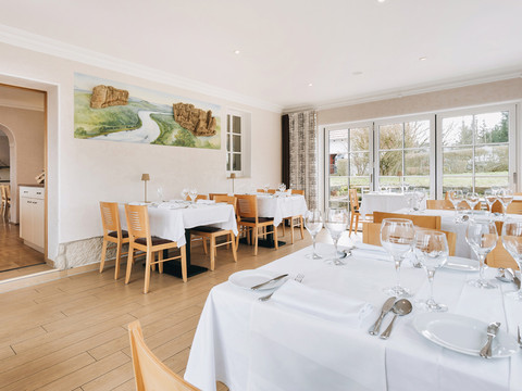 Helles Restaurant mit Holzboden, gedeckten Tischen und großen Fenstern mit Blick ins Grüne; an der Wand hängt ein Landschaftsbild.