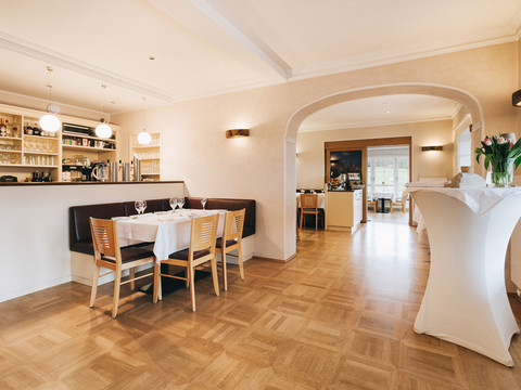 Helles Restaurant mit Holzboden, gedecktem Tisch und Stühlen, Barbereich im Hintergrund, weiße Tischdecke, Tulpenvase auf Stehtisch.