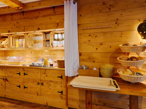 Ontbijtbuffet in een rustieke kamer met houten muren, gevuld met broodmandjes, sap, servies en bestek, verlicht door een warme lamp.