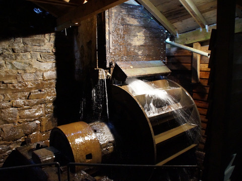 Wasserrad innen Oelchenshammer Ein hölzernes Wasserrad dreht sich in einer alten Mühle, umgeben von Steinwänden und Holzstruktur.