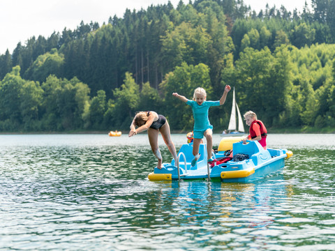 Freizeitcamp Aggertalsperre Ein Pedalboot auf einem See, mit Kindern die lachend ins Wasser springen; umgeben von Wald.