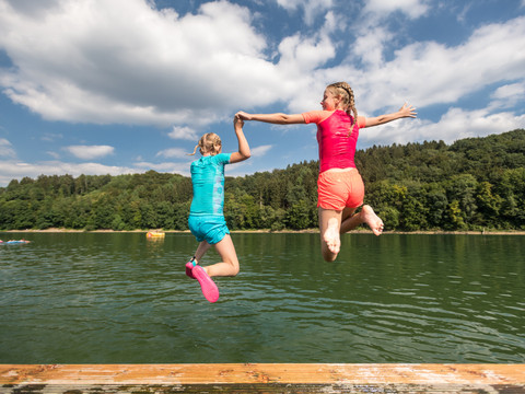 Freizeitcamp Aggertalsperre Zwei Mädchen springen Hand in Hand von einem Steg in einen ruhigen, von Bäumen gesäumten See.