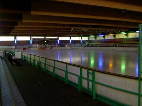 Mediendatei Leere Eissporthalle mit Holzbalkendecke, bunten Lichtern und grünem Geländer.