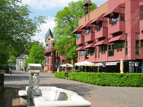 Bergischer Löwe Moderner Gebäudekomplex mit roten Fassaden neben historischem Turm und Springbrunnen bei sonnigem Wetter.