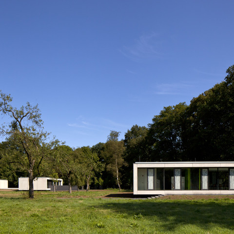 Gut Eichthal Zwei moderne, rechteckige Gebäude mit großen Glasfronten stehen auf einer grünen Wiese vor einem Wald.