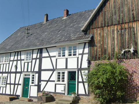 Fachwerkhäuser im Dorf Nümbrecht-Benroth <p>Fachwerkhaus mit grünem Eingang, weißen Wänden und schwarzen Balken neben Scheune bei klarem Himmel.</p>