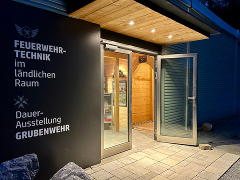Feuerwehrmuseum auf dem Kalkofenareal in Borna Eingang zur Dauerausstellung "Feuerwehrtechnik im ländlichen Raum" bei Dämmerung, mit offener Glastür und beleuchtetem Holzinterieur.Entrance to the permanent exhibition "Firefighting technology in rural areas" at dusk, with open glass door and illuminated wooden interior.Vstup do stálé expozice "Hasičská technika na venkově" za soumraku, s otevřenými skleněnými dveřmi a osvětleným dřevěným interiérem.Wejście na wystawę stałą "Technika pożarnicza na obszarach wiejskich" o zmierzchu, z otwartymi szklanymi drzwiami i oświetlonym drewnianym wnętrzem.Ingang van de permanente tentoonstelling "Brandweertechnologie in landelijke gebieden" bij schemering, met open glazen deur en verlicht houten interieur.Ingresso della mostra permanente "Tecnologia antincendio nelle aree rurali" al tramonto, con porta a vetri aperta e interni in legno illuminati.