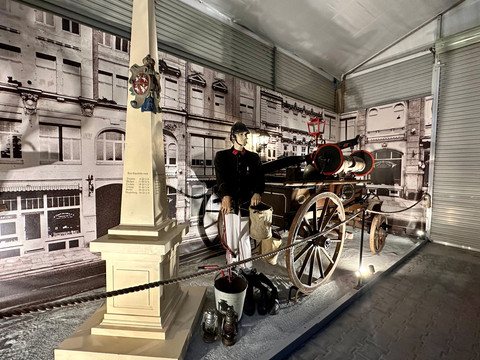 Einblick ins Feuerwehrmuseum Historische Feuerwehrszene mit einer Puppe in Uniform neben einer alten Feuerwehrkutsche, umgeben von Laternen und einem Obelisken.