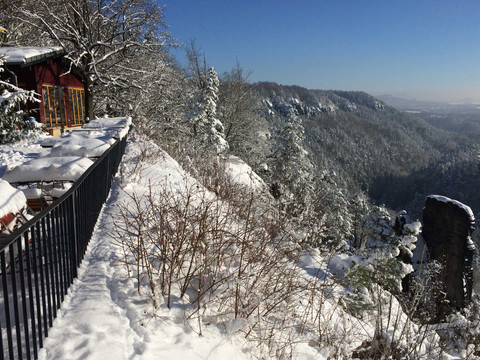 Verschneite Landschaft mit einem roten Gebäude links, Tischen unter Schnee und einem Blick auf bewaldete Hügel unter klarem, blauem Himmel.