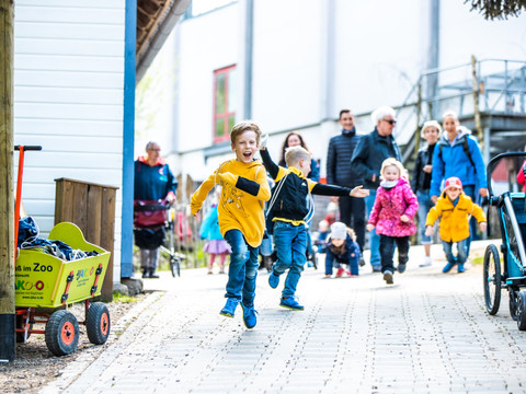 Affen- und Vogelpark Kinder laufen lachend auf einem gepflasterten Weg, gefolgt von Erwachsenen in Freizeitkleidung.