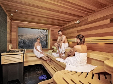 Wellness-Oase im Berghotel Bastei Drei Personen in Handtüchern sitzen in einer Holzsauna mit großem Fenster, das eine verschneite Landschaft zeigt. Entspannte Atmosphäre.