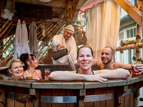 Winterdorf Schmilka Vier Personen entspannen in einem Holzbottich, lachen und trinken Bier; ein Mann gießt Wasser nach. Die Atmosphäre wirkt gemütlich und fröhlich.