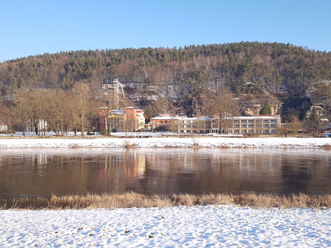 Sendig's wintertuin in het Parkhotel Flussufer im Winter mit schneebedecktem Boden, dahinter bunte Gebäude und ein bewaldeter Hügel unter klarem, blauem Himmel.