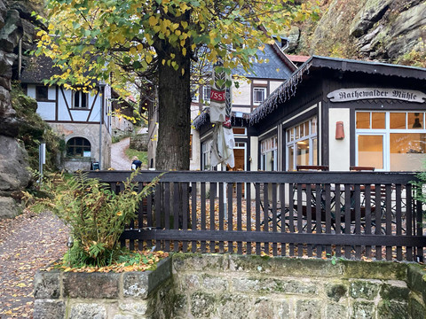 Rathewalder Mühle Fachwerkhaus mit der Aufschrift "Rathewalder Mühle", umgeben von herbstlichen Bäumen und Felsen, mit einem Weg im Vordergrund.