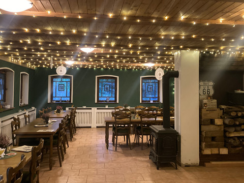 Gemütlicher Raum mit Holztischen und -stühlen, Lichterketten an der Decke, grüne Wände, Kaminofen und gestapeltes Holz; Route 66 Schild an der Wand.Cozy room with wooden tables and chairs, fairy lights on the ceiling, green walls, wood-burning stove and stacked wood; Route 66 sign on the wall.Útulná místnost s dřevěnými stoly a židlemi, pohádkovými světly na stropě, zelenými stěnami, kamny na dřevo a naskládaným dřevem; na stěně nápis Route 66.Przytulny pokój z drewnianymi stołami i krzesłami, lampkami na suficie, zielonymi ścianami, piecem opalanym drewnem i stosem drewna; znak Route 66 na ścianie.Gezellige kamer met houten tafels en stoelen, feeërieke lichtjes aan het plafond, groene muren, houtkachel en gestapeld hout; Route 66-bord aan de muur.Locale accogliente con tavoli e sedie in legno, luci fiabesche sul soffitto, pareti verdi, stufa a legna e legna accatastata; insegna della Route 66 alla parete.