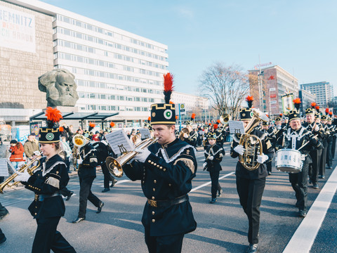 Bergparade vorbei am Karl-Marx-Kopf. © Ernesto Uhlmann_CWE.jpg Musikkapelle in traditioneller Uniform marschiert vor dem Karl-Marx-Denkmal in einer Stadt.
