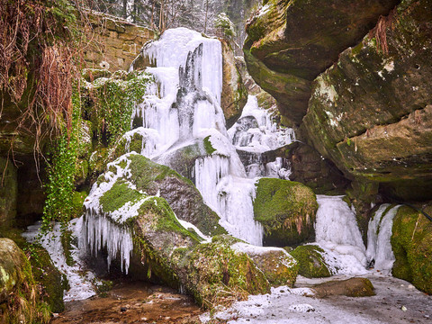 Vereister Wasserfall zwischen moosbewachsenen Felsen, umgeben von winterlicher Vegetation und einer ruhigen, frostigen Atmosphäre.