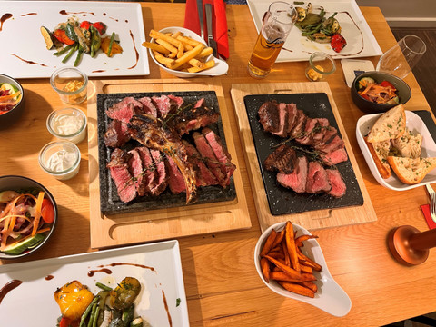 Haus Olpe Ein gedeckter Tisch mit verschiedenen Grillsteaks, Beilagen wie Pommes, Gemüse und einem Glas Bier.