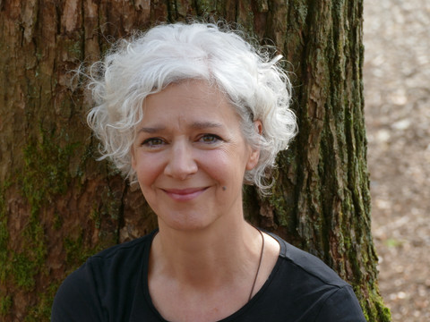 Porträt Ältere Frau mit grauen Locken, steht lächelnd vor einem Baum mit Moos, entspannter Ausdruck.