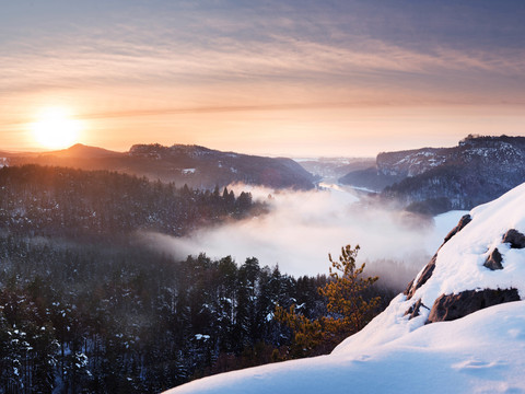 Winterliche Landschaft mit schneebedeckten Bergen und Wäldern im Vordergrund, Nebel über dem Tal und einem Sonnenuntergang am Horizont.