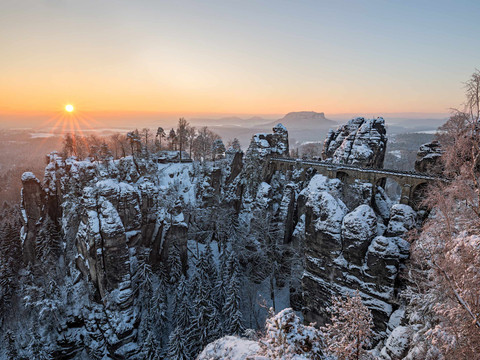 Verschneite Felsformationen und eine Steinbrücke im Sonnenaufgang, umgeben von schneebedeckten Bäumen, schaffen eine friedliche Winterlandschaft.
