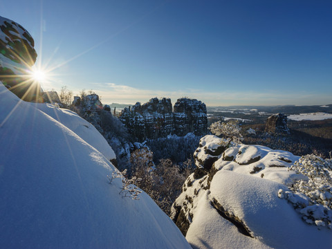 Verschneite Schrammsteine bei Sonnenaufgang, die Sonne strahlt über schneebedeckte Hügel, klare blaue Himmel im Hintergrund.