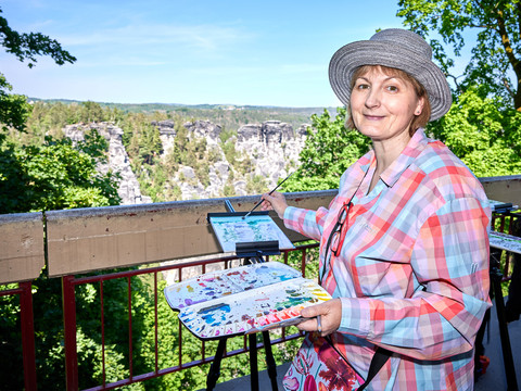 Malkurs an der Bastei mit Andrea Moliére Eine Frau mit Hut malt auf einer Staffelei in einer grünen, felsigen Landschaft. Sie trägt ein bunt kariertes Hemd und hält eine Farbpalette.