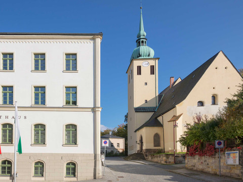 Kostel svatého Petra a Pavla Sebnitz Weißes Rathausgebäude mit grünen Fensterrahmen und daneben eine Kirche mit grünem Turm vor blauem Himmel.