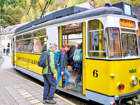 Die Kirnitzschtalbahn im Nationalpark Sächsische Schweiz Menschen steigen in eine gelbe Straßenbahn der Kirnitzschtalbahn ein, umgeben von grüner Landschaft und Gebäuden im Hintergrund.
