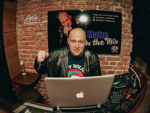 DJ mit Mikrofon und Laptop vor einer Ziegelwand, umgeben von Audioequipment in einer Bar.