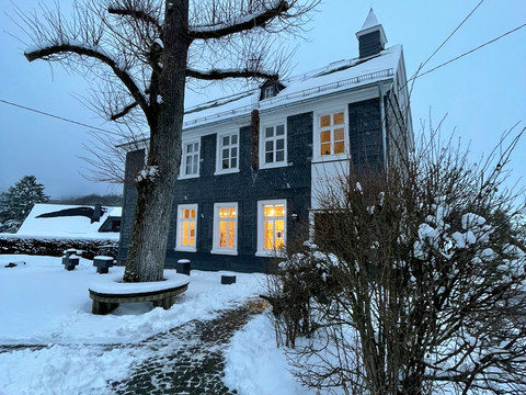 Kunst Kabinett Hespert Ein zweistöckiges Haus bei Dämmerung, eingeschneit, mit leuchtenden Fenstern und einem großen Baum.