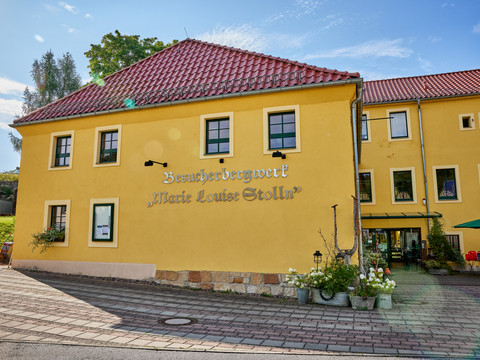 Scheidehaus am "Marie Louise Stolln" Gelbes Gebäude in traditionellem Stil mit rotem Ziegeldach, umgeben von blühenden Blumen und Gehweg.
