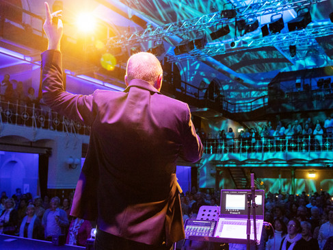 Een omroeper staat voor een publiek in een verlichte evenementenzaal.