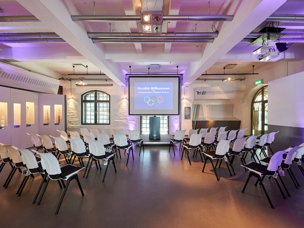 Olympia Lounge (21).jpg Ein moderner Konferenzraum mit weißen Stühlen, einem Projektor und beleuchteten Wänden.</p>A modern conference room with white chairs, a projector and illuminated walls.</p> <p