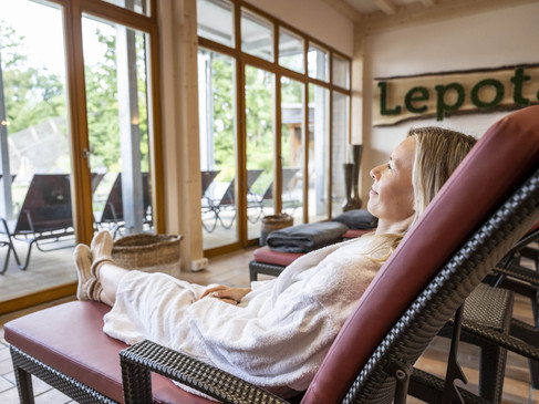 Eine Frau entspannt in einem Spa-Bereich auf einer Liege, umgeben von großen Fenstern mit Gartenblick.