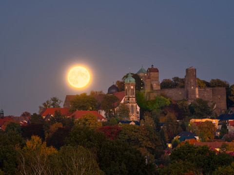 Burg Stolpen Vollmond über der Burg Stolpen mit Türmen und roten Dächern, umgeben von herbstlichen Bäumen; ruhige, stimmungsvolle Abenddämmerung.