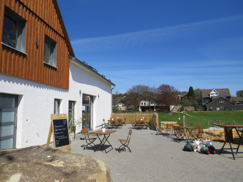 Hofladen Außenbereich  Sonnige Terrasse mit Holztischen und -stühlen vor einem Café mit weißer Fassade, ländliche Umgebung.