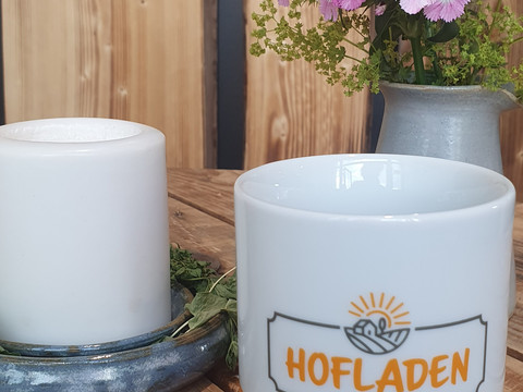 Hofladen Tasse Weiße Tasse mit "Hofladen Ülfetal"-Logo, daneben Kerze und pinke Blumen auf Holztisch.