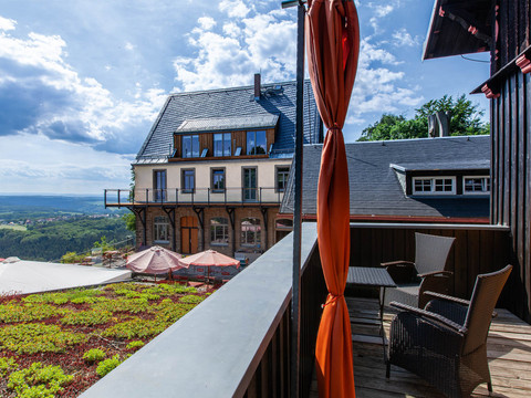 Blick von einer Terrasse mit Rattanmöbeln auf ein großes Gebäude mit Schieferdach, umgeben von grüner Landschaft und blauem Himmel.